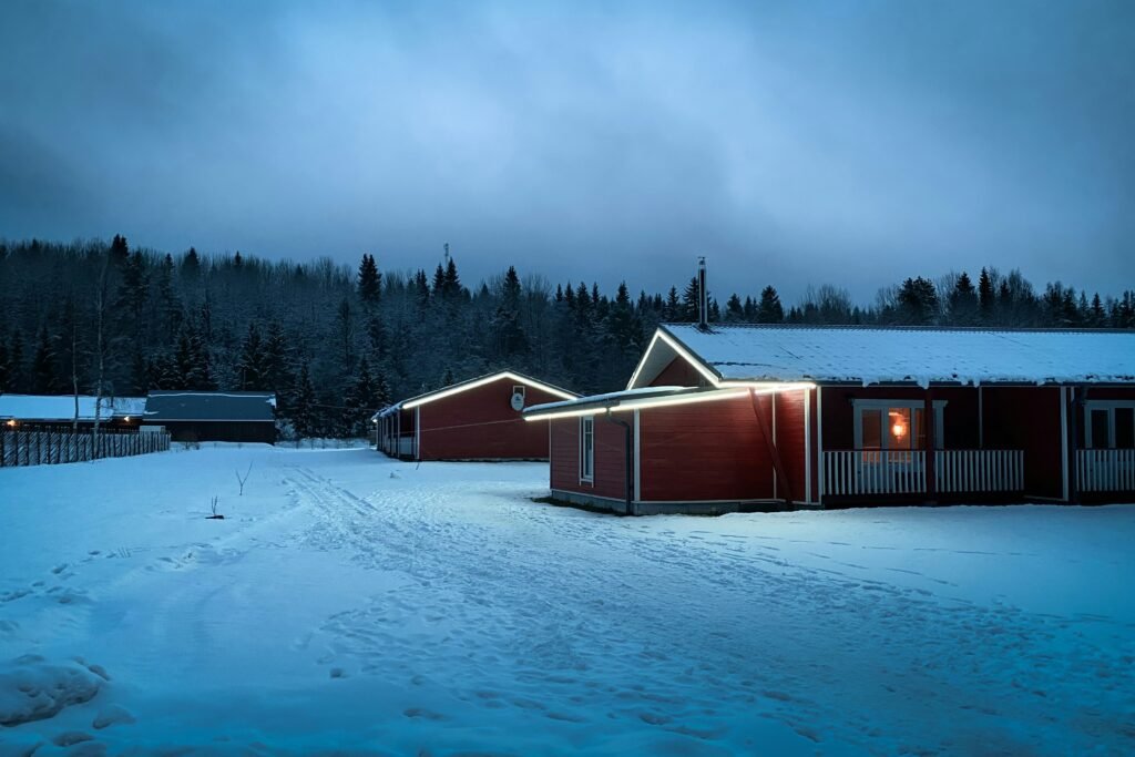 A peaceful winter scene at dusk featuring red wooden cabins with white trim, softly illuminated by exterior lights. The ground and rooftops are covered in snow, and the cabins are surrounded by a snowy forest with tall evergreen trees under a cloudy blue sky.