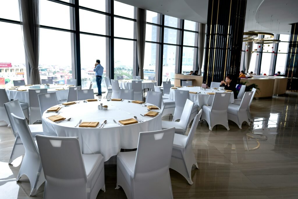 Modern upscale restaurant with large glass windows, round tables covered in white tablecloths, and neatly arranged white chairs; a man stands by the window on the phone while a few guests dine in the background.
